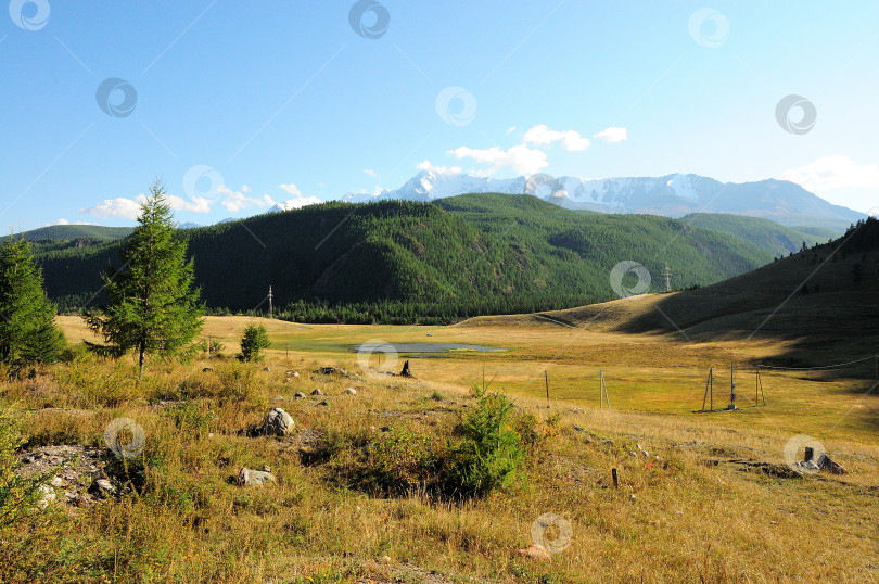 Скачать Вид с вершины на небольшую долину с небольшим озером и столбами электропередач на фоне высоких гор с заснеженными вершинами. фотосток Ozero
