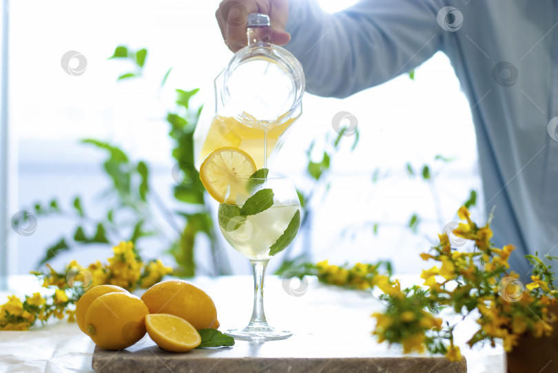 Скачать Вручную наливаем желтый лимонад со льдом в бокалы, украшенные ломтиками лимона, и на мраморный стол на естественном фоне фотосток Ozero