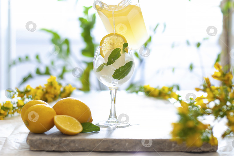 Скачать Разлив желтого лимонада со льдом по бокалам с украшением ломтиками лимона на мраморном столе на естественном фоне. Вытекающий сок фотосток Ozero