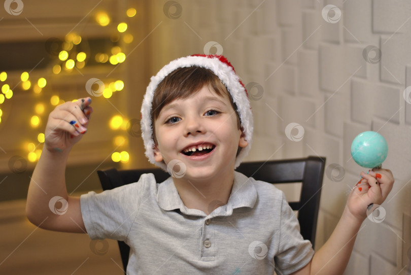 Скачать Веселый, счастливый, улыбающийся маленький мальчик раскрашивает рождественские шары в красно-белой пушистой шапочке. Праздничные украшения ручной работы фотосток Ozero