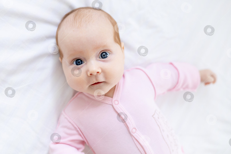Скачать портрет маленькой новорожденной девочки с голубыми глазами крупным планом лежит дома на белой хлопчатобумажной кровати и улыбается фотосток Ozero