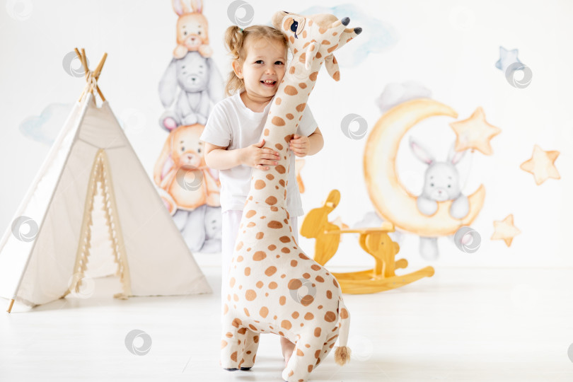 Скачать маленькая девочка-блондинка играет в детской комнате, обнимает большого игрушечного жирафа, измеряет свой рост, детский досуг, детский сад фотосток Ozero