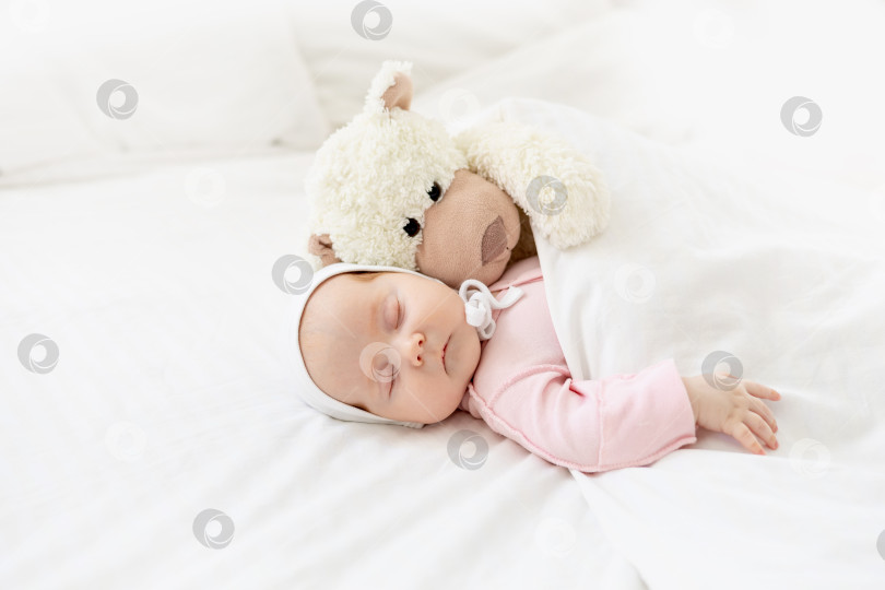 Скачать крупным планом портрет маленькой новорожденной девочки, спящей дома на белой хлопчатобумажной кровати, обнимающей плюшевого зайца под одеялом в чепчике фотосток Ozero