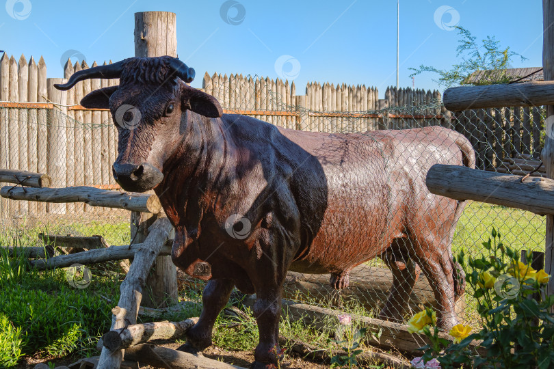 Скачать Декоративный бык выглядывает из ограждения на фоне изгорди и деревянного забора из бревен. фотосток Ozero