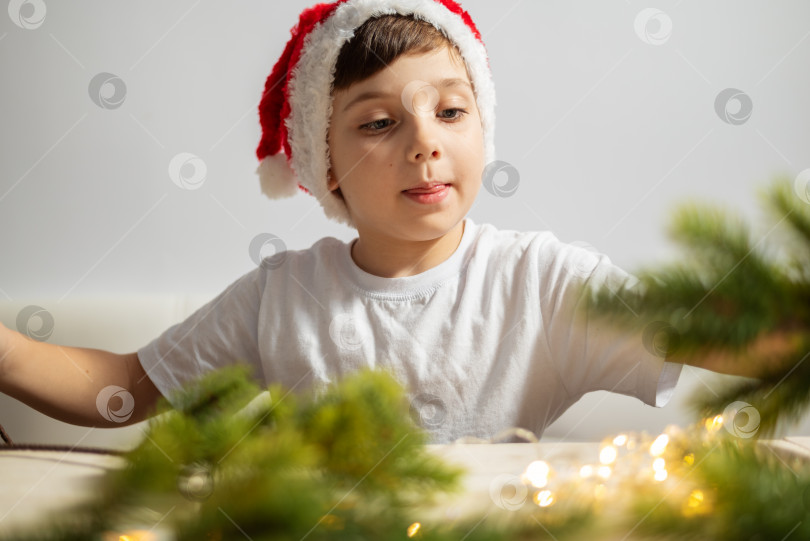 Скачать Милый маленький мальчик в рождественской шляпе Санты, украшающий дом еловыми ветками фотосток Ozero