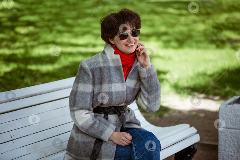 Скачать Молодая жизнерадостная женщина разговаривает по телефону, сидя на скамейке фотосток Ozero