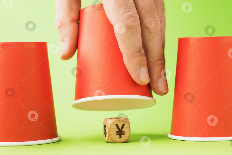 Скачать Поднятый бумажный стаканчик и деревянный кубик с символом юаня внизу, концепция азартных игр в наперсток фотосток Ozero