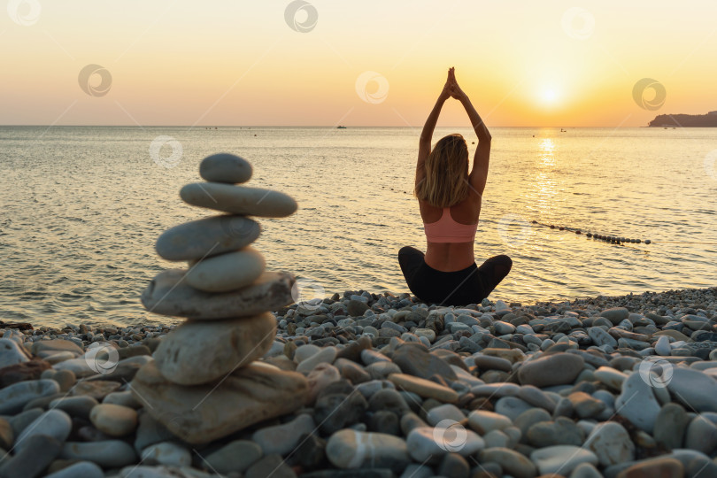 Скачать Женщина в спортивной одежде, практикующая йогу, занимается медитацией, сидя в позе лотоса на берегу моря, рядом с ней выложена пирамида из камней фотосток Ozero