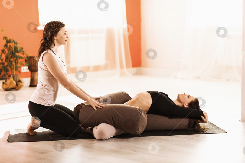 Скачать Две женщины в спортивной одежде, практикующие йогу, выполняют упражнение Супта баддха конасана с валиками на гимнастическом коврике в студии фотосток Ozero