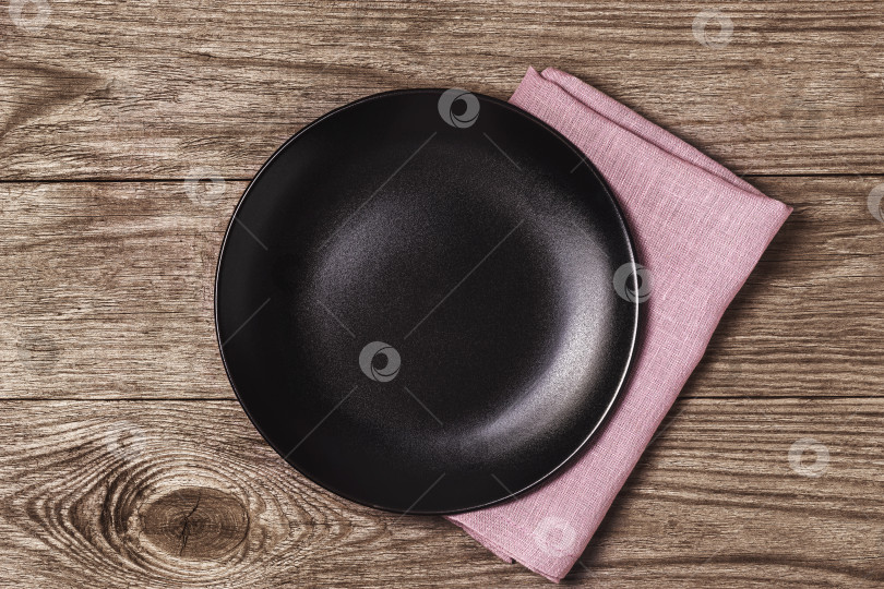 Скачать Пустая черная керамическая тарелка с фиолетовой салфеткой на деревянном столе, вид сверху. Пищевой фон фотосток Ozero