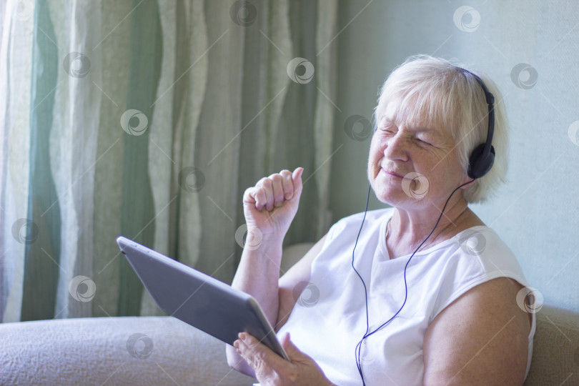 Скачать Улыбающаяся пожилая женщина со светлыми волосами совершает видеозвонок дома, используя цифровой планшет и наушники. удаленная связь. технологии. фотосток Ozero