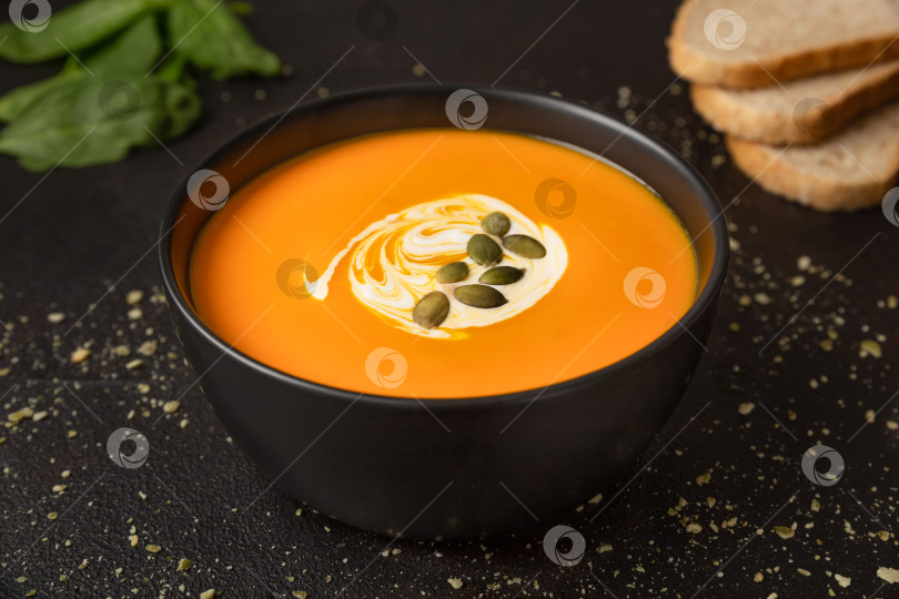 Скачать Вегетарианский домашний тыквенно-морковный суп со сливками и тыквенными семечками в черной миске на темном фоне крупным планом фотосток Ozero