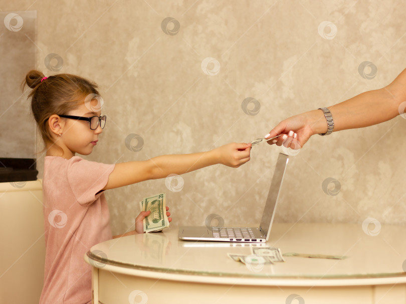Скачать девочка 5 лет в розовом платье и очках за столом с компьютером протягивает руку с банкнотами в долларах. фотосток Ozero