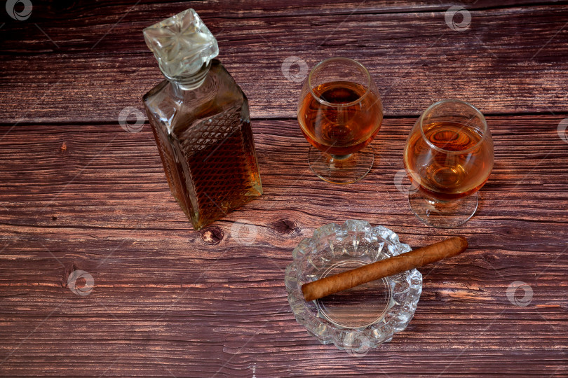 Скачать Украшенный хрусталем графин и два бокала с коньяком на деревянном столе, рядом с кубинской сигарой в стеклянной пепельнице. фотосток Ozero