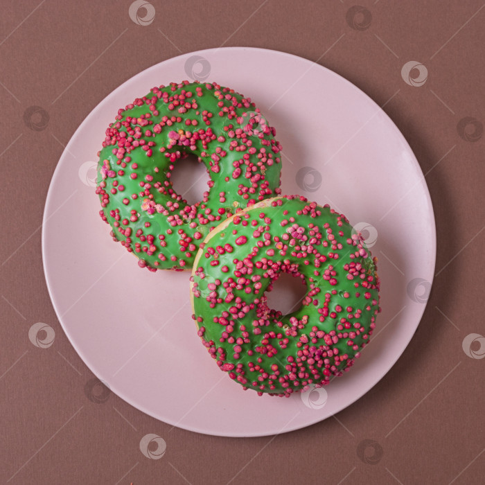 Скачать Два зеленых пончика с красной и розовой посыпкой на розовом блюдце на коричневом фоне. Вид сверху фотосток Ozero
