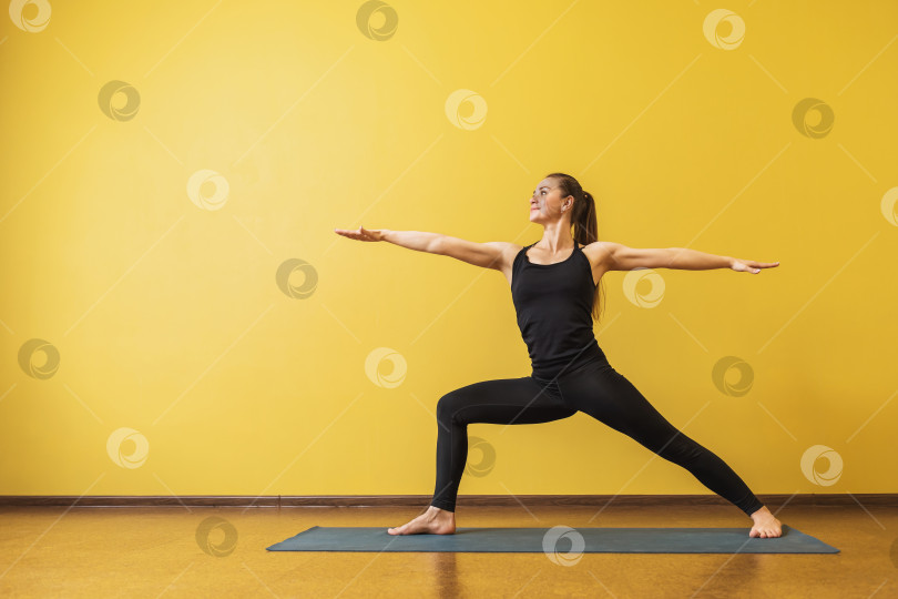 Скачать Привлекательная женщина в черной спортивной одежде, практикующая йогу, выполняет упражнение Вирабхадрасана, поза воина 2, на фоне желтой стены с местом для копирования фотосток Ozero