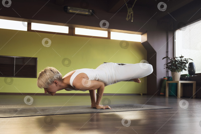 Скачать Блондинка в белой спортивной одежде, практикующая йогу, выполняет упражнение Падма Маюрасана, позу павлина, асану равновесия на коврике в студии фотосток Ozero