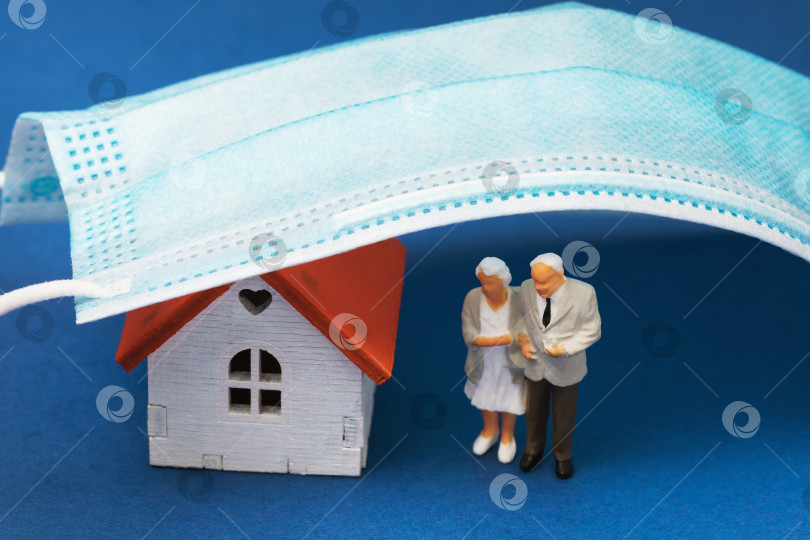 Скачать Пара пенсионеров возле дома и медицинская маска на синем фоне, концепция о необходимости оставаться дома для пожилых людей, игрушечные человечки из пластика фотосток Ozero