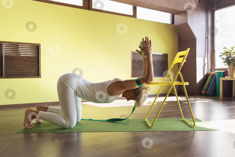 Скачать Женщина, практикующая йогатерапию, выполняет упражнение на растяжку мышц позвоночника с помощью стула и ремней, тренируется в студии на коврике фотосток Ozero