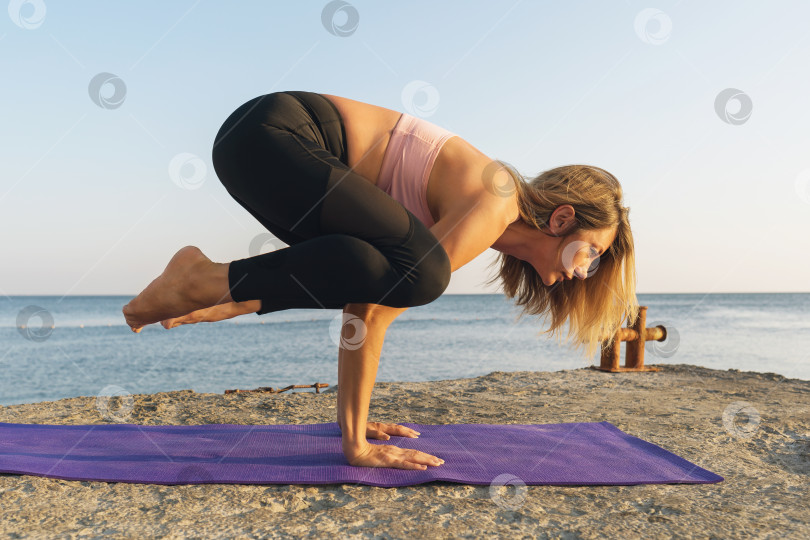 Скачать Женщина в спортивной одежде, практикующая йогу на берегу моря, выполняет упражнение какасана, поза вороны фотосток Ozero