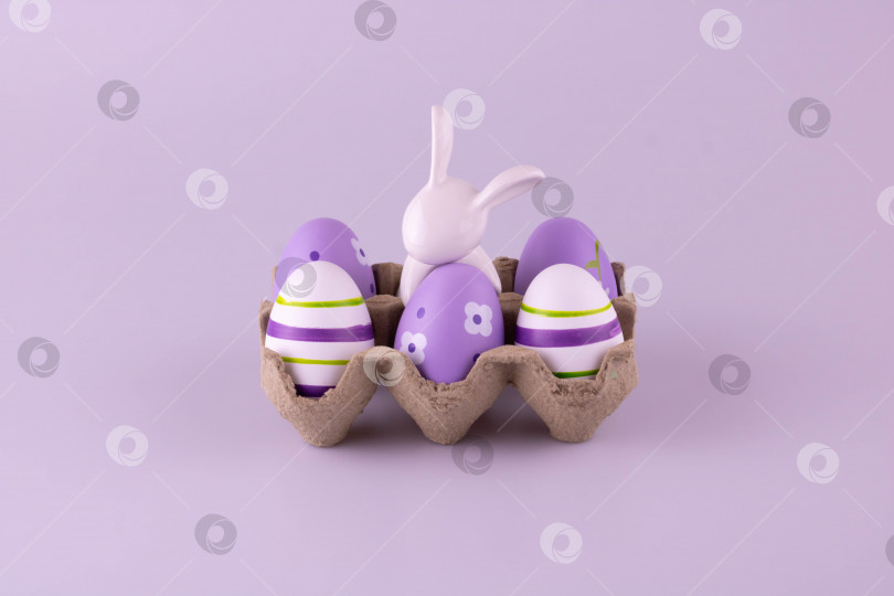 Скачать Белый керамический кролик в коробке для яиц, рядом с ним лежат пасхальные яйца. фотосток Ozero
