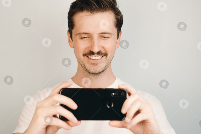 Скачать Улыбающийся молодой человек смотрит на телефон в помещении. Портрет усатого мужчины в футболке, снимающий на камеру мобильного телефона фотосток Ozero