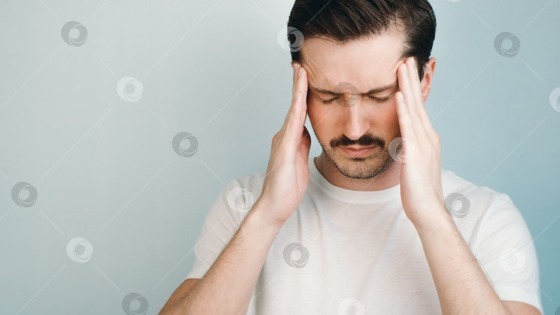 Скачать Концепция головной боли, усталости. Мужчина в белой футболке держится руками за голову в помещении, массируя виски. фотосток Ozero