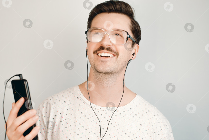 Скачать Молодой человек в больших очках улыбается и слушает музыку по телефону в помещении, портрет крупным планом. фотосток Ozero
