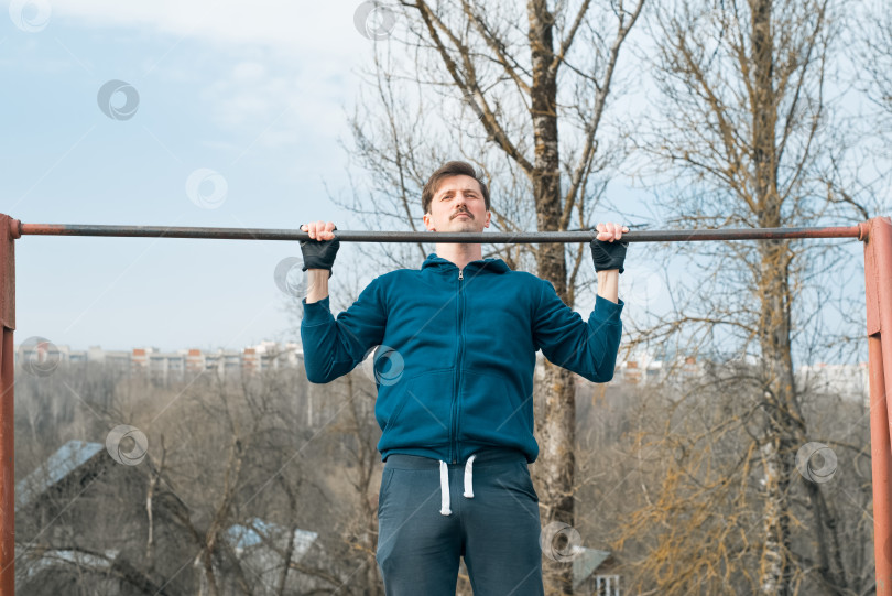 Скачать Молодой человек в спортивной одежде тренируется на улице, подтягиваясь на турнике. Силовая тренировка на свежем воздухе фотосток Ozero