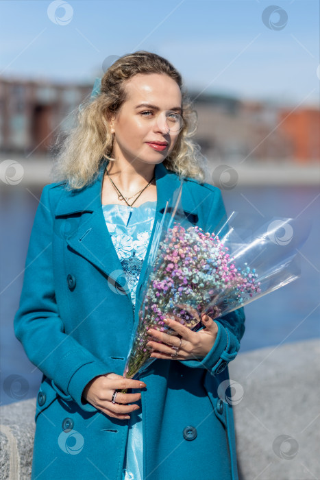 Скачать Молодая блондинка в синем пальто стоит с букетом разноцветных гипсофил на набережной в парке. Концепция весеннего праздника - 8 марта, Пасха, Женский день фотосток Ozero