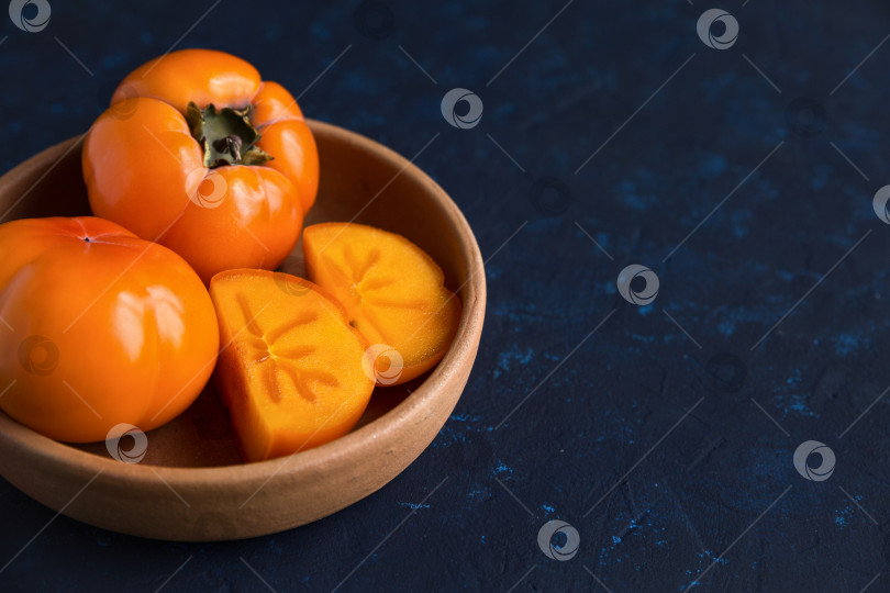 Скачать Оранжевая сочная спелая хурма лежит в глиняной тарелке на темно-синем фоне. Хурму разрезают на две части. фотосток Ozero
