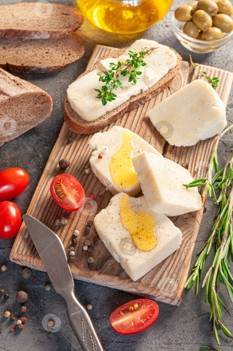 Скачать Адыгейский сыр нарезают кусочками. Сыр выложите на деревянную доску. Подается с тимьяном, розмарином, специями, помидорами черри и оливковым маслом. На заднем плане - оливки, ржаной хлеб и оливковое масло. Фон из серого бетона. Вид сверху. фотосток Ozero