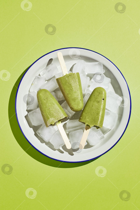 Скачать Замороженное домашнее фисташковое эскимо в миске со льдом на зеленом фоне. Освежающее фруктовое мороженое, замороженный зеленый сок на палочке. Вид сверху, место для копирования фотосток Ozero