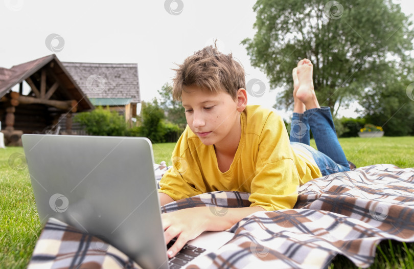 Скачать Четырнадцатилетний мальчик с ноутбуком сидит в парке. Школьник печатает на компьютере на открытом воздухе. Молодой парень учится онлайн за компьютером. Мальчик ведет онлайн-урок, находясь на улице. фотосток Ozero
