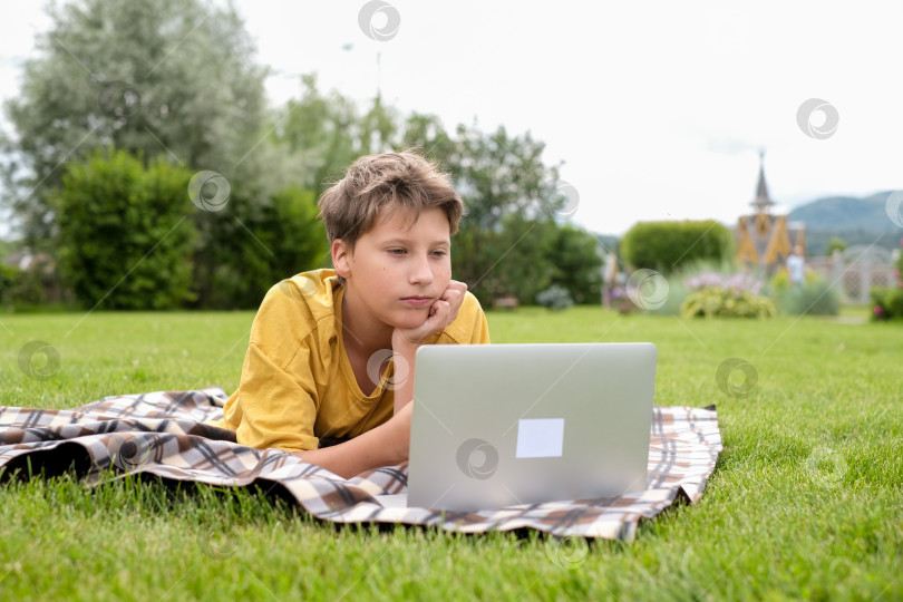 Скачать Четырнадцатилетний мальчик с ноутбуком сидит в парке. Школьник печатает на компьютере на открытом воздухе. Молодой парень учится онлайн за компьютером. Мальчик ведет онлайн-урок, находясь на улице. фотосток Ozero