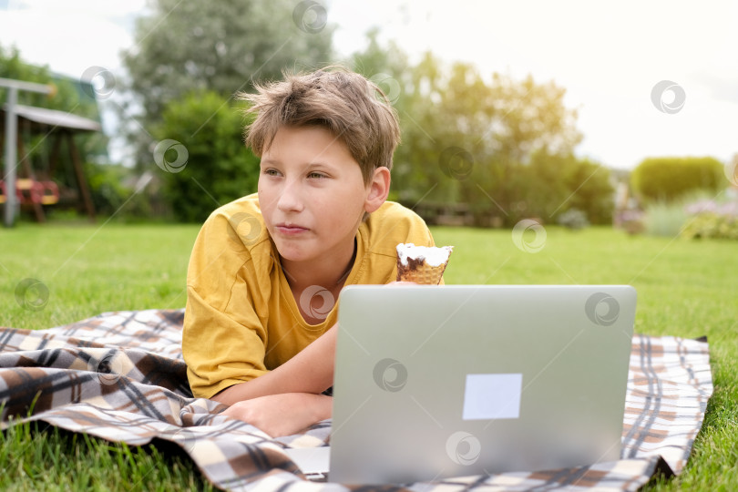 Скачать Мальчик-подросток в желтой футболке подстилает траву, ест мороженое, улыбается и печатает на ноутбуке в парке. Школьник с компьютером на улице. Молодой мальчик учится или играет в компьютерные игры онлайн за компьютером. Мальчик ведет онлайн-урок, находясь на улице. фотосток Ozero