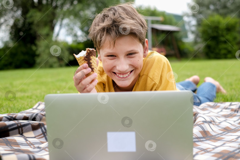 Скачать Мальчик-подросток в желтой футболке подстилает траву, ест мороженое, улыбается и печатает на ноутбуке в парке. Школьник с компьютером на улице. Молодой мальчик учится или играет в компьютерные игры онлайн за компьютером. Мальчик ведет онлайн-урок, находясь на улице. фотосток Ozero