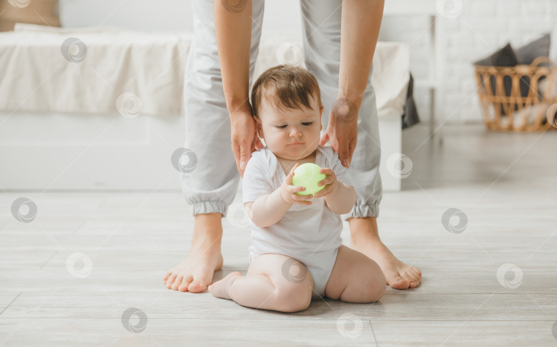 Скачать Милый маленький малыш 6 месяцев от роду сидит на полу рядом с мамиными ногами и улыбается, мама держит его за руки фотосток Ozero