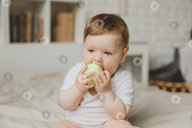 Скачать Малыш в возрасте 6 месяцев грызет мяч, держа его руками, сидя на кровати. прорезывание зубов. фотосток Ozero