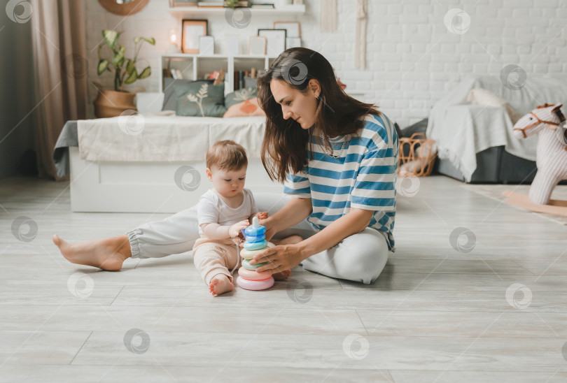 Скачать Красивая молодая мама играет и учит своего 6-месячного ребенка на полу в гостиной. мама и малыш играют с игрушками фотосток Ozero