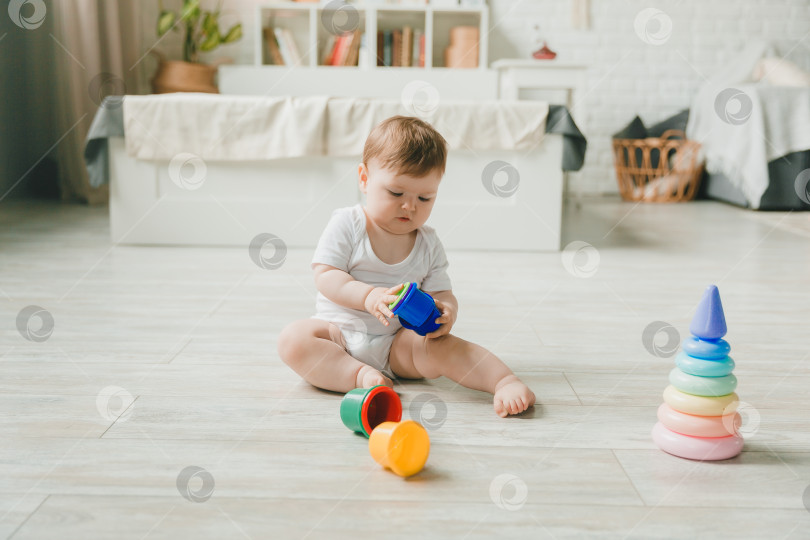 Скачать малыш 6-9 месяцев играет с разноцветной радужной игрушечной пирамидкой, сидя в белой солнечной спальне. Игрушки для маленьких детей. Детский интерьер. Ребенок с развивающей игрушкой. фотосток Ozero