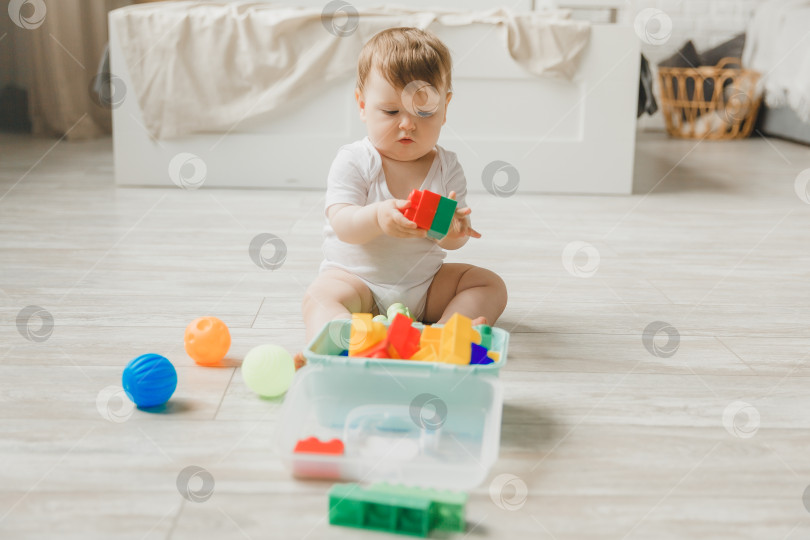 Скачать малыш 6-9 месяцев играет с разноцветной радужной игрушечной пирамидкой, сидя в белой солнечной спальне. Игрушки для маленьких детей. Детский интерьер. Ребенок с развивающей игрушкой. фотосток Ozero