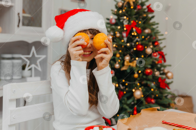 Скачать Счастливая кавказская девушка в красной шапочке Санты держит в руке мандарины. На заднем плане изображена рождественская елка. фотосток Ozero
