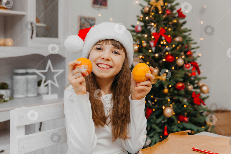 Скачать Счастливая кавказская девушка в красной шапочке Санты держит в руке мандарины. На заднем плане изображена рождественская елка. фотосток Ozero