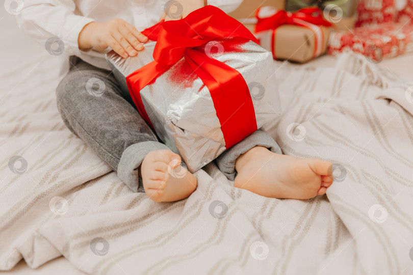 Скачать Счастливый ребенок в шапке Санты сидит на полу возле рождественской елки и держит в руках коробку с подарком. голые пятки крупным планом фотосток Ozero