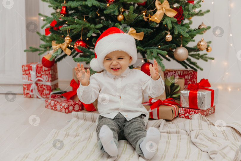 Скачать очаровательный маленький мальчик в новогодней шапочке сидит на одеяле перед рождественской елкой. фотосток Ozero