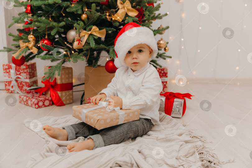 Скачать Счастливый малыш в шапке Санты сидит на полу возле рождественской елки и держит в руках коробку с подарком. фотосток Ozero