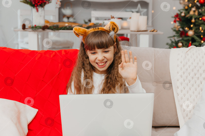 Скачать Маленькая девочка в шапочке Санта-Клауса общается по видеосвязи через ноутбук, сидя на диване в гостиной. фотосток Ozero