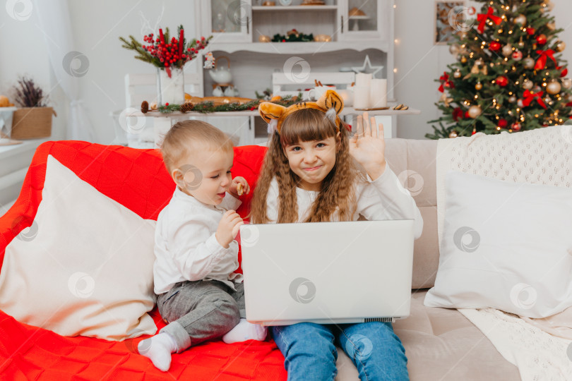 Скачать Маленькая девочка в шапочке Санта-Клауса и ее брат ведут видеосвязь через ноутбук, сидя на диване в гостиной. фотосток Ozero