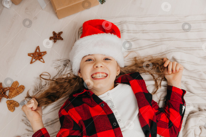 Скачать маленькая девочка в шапочке Санта-Клауса лежит дома возле рождественской елки. вид сверху. Рождество. фотосток Ozero